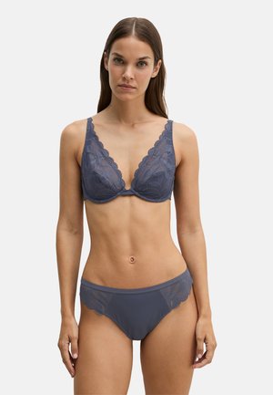 Vrouw met lang bruin haar die een bijpassende donkergrijze kanten bh en slip draagt, staand tegen een effen lichte achtergrond.