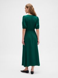 Woman facing away wearing a dark green midi dress with puff sleeves and black flat shoes on a plain white background.