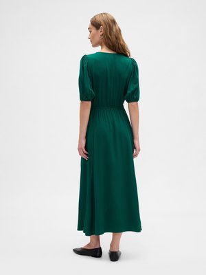 Woman facing away wearing a dark green midi dress with puff sleeves and black flat shoes on a plain white background.