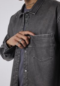 Giacca in denim grigio con bottoni a scatto, tasca frontale e cuciture bianche a contrasto. La mano del modello sta raggiungendo la tasca.