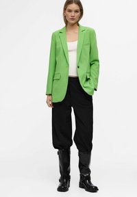 Groene blazer met scherpe revers, twee voorzakken en één knoopsluiting, gecombineerd met zwarte baggy broek en kniehoge zwarte laarzen.