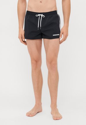 Personne torse nu portant un maillot de bain court noir avec un cordon blanc et des rayures sur les côtés, debout pieds nus sur un fond blanc.