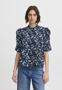 Navy blue blouse with white floral pattern, short puffed sleeves, buttoned front, and straight cut design. Paired with light blue denim jeans.