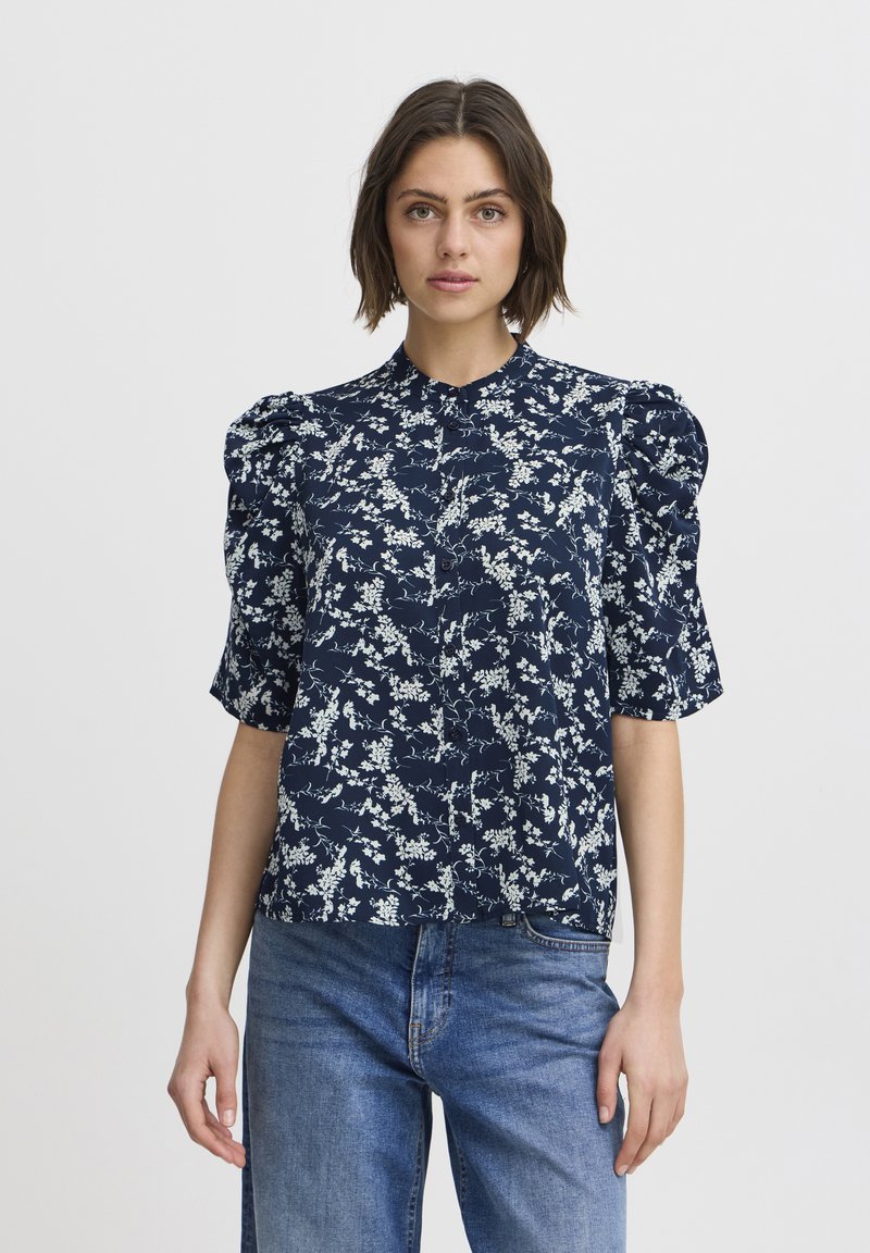 Navy blue blouse with white floral pattern, short puffed sleeves, buttoned front, and straight cut design. Paired with light blue denim jeans.