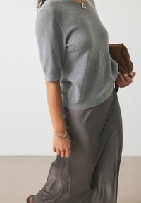 Maglione grigio a maniche corte con scollatura rotonda, abbinato a una gonna fluida grigio scuro. Tiene in mano una borsa a tracolla marrone e indossa un bracciale argentato.