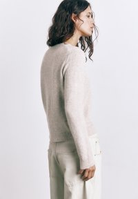 Mladá žena s kučeravými vlasmi, oblečená v béžovom svetri a svetlých nohaviciach, stojaca v profile na jednoduchom bielom pozadí.