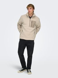 Pull polaire beige avec col montant, poche zippée, deux poches latérales et finition texturée, associé à un jean noir et des baskets.