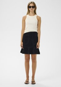 Top sans manches côtelé crème associé à une jupe évasée noire avec un motif texturé. Le modèle porte des lunettes de soleil noires et des sandales plates.