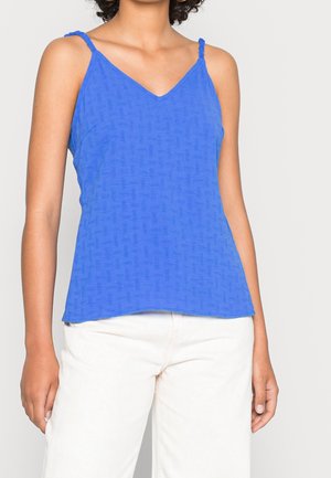 Femme portant un haut bleu sans manches à col en V avec des bretelles tressées et un pantalon blanc, debout devant un fond clair uni.