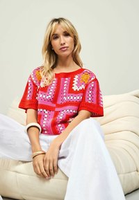 Haken-stijl trui met korte mouwen in rood, roze en wit met geometrische patronen. Model zit tegen een textuurachtige crèmekleurige achtergrond, gekleed in witte broeken.
