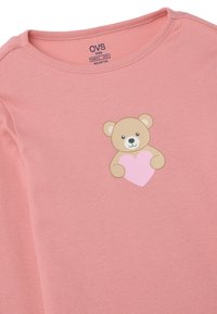 Camisa de manga larga de color rosa, de algodón orgánico, con un gráfico de un oso sosteniendo un corazón en el pecho. La etiqueta dice "OVS kids."