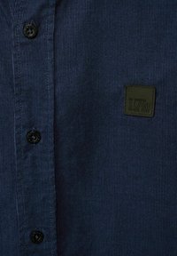 Tmavě modrá manšestrová košile s černými knoflíky a zelenou nášivkou s logem, která má vroubkovanou texturu a klasický límeček.