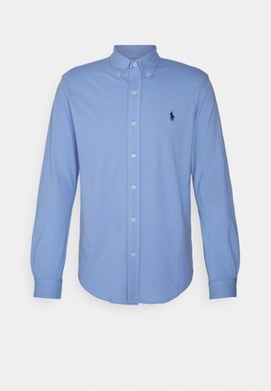 Camisa azul claro de manga larga con botones, cuello abotonado y pequeño logo oscuro en el pecho izquierdo.