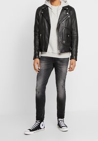 Chaqueta de cuero negra de motociclista sobre una sudadera gris, combinada con jeans gris ajustados y zapatillas negras de caña alta con acentos blancos.