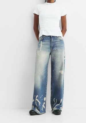 Personne portant un jean taille haute à jambes larges bleu délavé avec des éclaboussures de peinture blanche et un haut blanc ajusté à manches courtes, debout contre un mur.