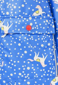 Blauer Stoff mit weißen und gelben Gänseblümchen und weißen Pferden mit rosa Mähnen, ausgestattet mit einer Tasche mit einem roten Druckknopf.