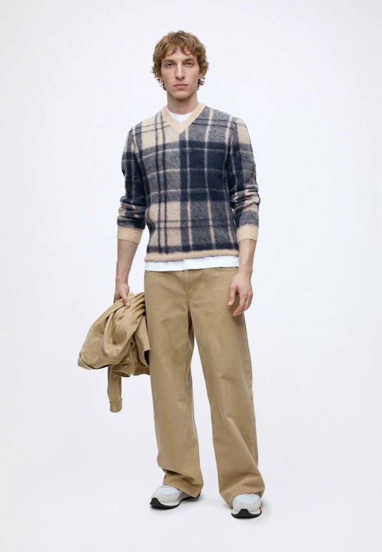 Jeune homme portant un pull beige et bleu marine à carreaux, un pantalon beige ample, des baskets blanches, tenant une veste beige dans une main.