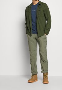 Camisa verde de manga larga sobre una camiseta azul, combinada con pantalones cargo verde claro y botas beige. Presenta bolsillos y un ajuste relajado.