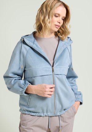 Donna con capelli biondi fino alle spalle, che indossa una felpa con cappuccio azzurro chiaro con cerniera e pantaloni beige, guarda verso il basso con una mano in tasca.