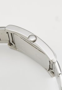 Stříbrné náramkové hodinky s kulatým pouzdrem, hladkým kovovým páskem a texturovanou korunou. Mají lesklý povrch a minimalistický design.
