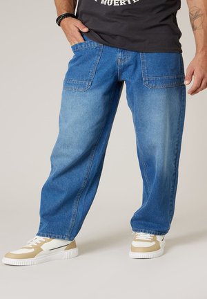 Homme portant un jean décontracté en denim bleu avec de grandes poches avant, des baskets blanches et beiges, et un t-shirt noir, debout avec une main dans la poche.