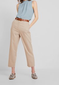 Top senza maniche azzurro chiaro con pieghe, pantaloni larghi beige e scarpe con motivo nelle tonalità di marrone e bianco. Una cintura marrone completa l'outfit.