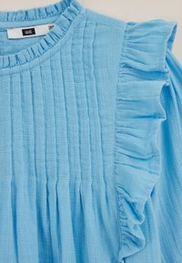 Svetlo modra bombažna majica z mehkimi teksturami, nabranim sprednjim delom in volanastimi rokavi. Okrogli izrez z nabranim zaključkom na ovratniku.