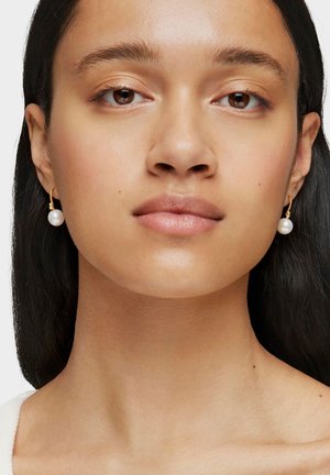 Jeune femme à la peau mate portant des boucles d'oreilles créoles dorées avec des perles blanches pendantes, vue du cou jusqu'au sommet de la tête sur un fond blanc.