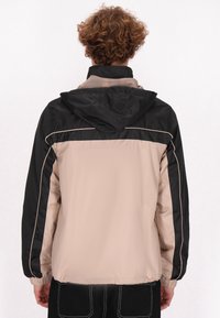 Lichtgewicht, met een capuchon, beige en zwarte jas, met contrasterende zijstrepen en elastische manchetten. Gladde textuur met een sportief ontwerp.