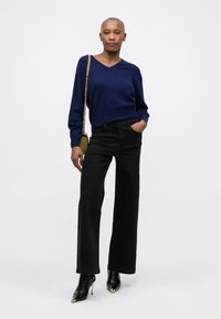Marineblauwe gebreide trui met een V-hals, gecombineerd met hoge zwarte wijde jeans en pointed zwarte enkellaarsjes met gouden accenten.