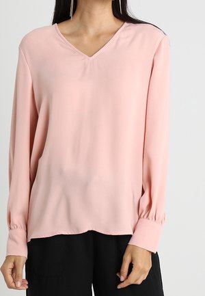 Femme portant une blouse rose clair à manches longues avec col en V et poignets à boutons, associée à un pantalon noir, sur un fond uni.