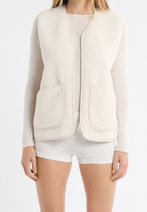 Femme portant un gilet sherpa crème sans manches avec des poches avant, sur un haut à manches longues transparent et un short blanc, debout devant un fond blanc.