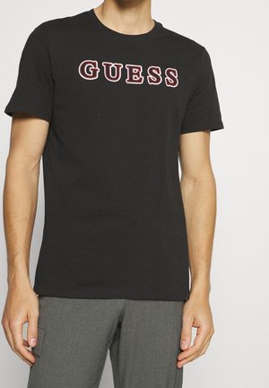 Mann trägt ein schwarzes T-Shirt mit "GUESS" in fetten weißen und roten Buchstaben auf der Brust und graue Hose mit Seitentasche.