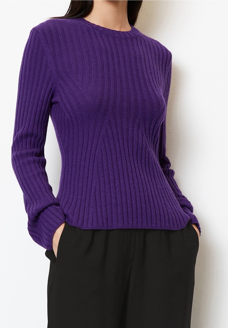Femme portant un pull ajusté violet en maille côtelée à manches longues et un pantalon noir avec poches, mains glissées dans les poches.