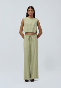 Top corto verde chiaro con ampi fori per le braccia, abbinato a pantaloni aperti coordinati. Il tessuto sembra liscio e leggero, con una vita regolabile.
