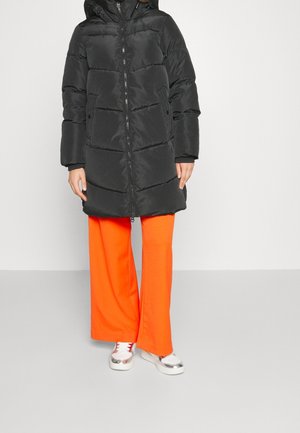 Persoon draagt een zwarte, doorgestikte, gewatteerde winterjas met capuchon, feloranje broek met wijde pijpen en witte sneakers met metallic details.