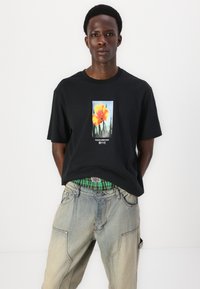 Tricou negru din bumbac, cu un design grafic floral în nuanțe de portocaliu și verde, asociat cu pantaloni din denim decolorat, cu detalii de cusătură.