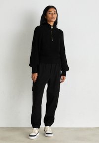 Černý pletený svetr s vysokým límcem a zlatým zipem, v kombinaci s černými cargo kalhotami a bílými teniskami s černými akcenty.