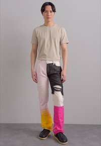 T-shirt beige abbinato a jeans patchwork multicolore con pannelli rosa, gialli e neri; dettagli strappati e calzature robuste.
