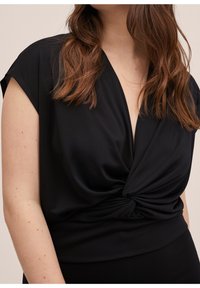 Femme portant un haut noir sans manches avec un col en V profond et un détail de nœud torsadé à la taille, cheveux bruns mi-longs visibles.