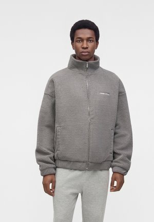 Un homme porte une veste polaire grise zippée à col montant et un pantalon de jogging assorti gris clair, sur un fond blanc uni.