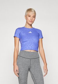 Lilla kortermet crop-top med et teksturert mønster, med rund hals og et hvitt Adidas-logo, parret med grå leggings.