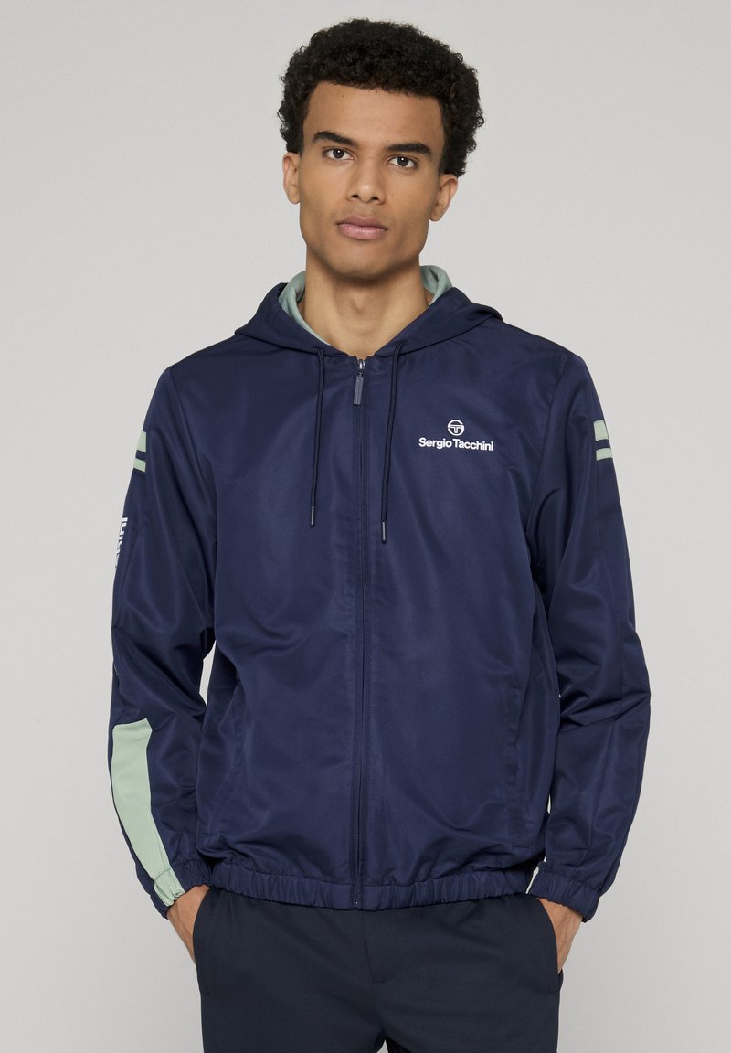 Jeune homme portant une veste à capuche zippée Sergio Tacchini bleu marine avec des accents verts clairs sur les manches, debout les mains dans les poches.