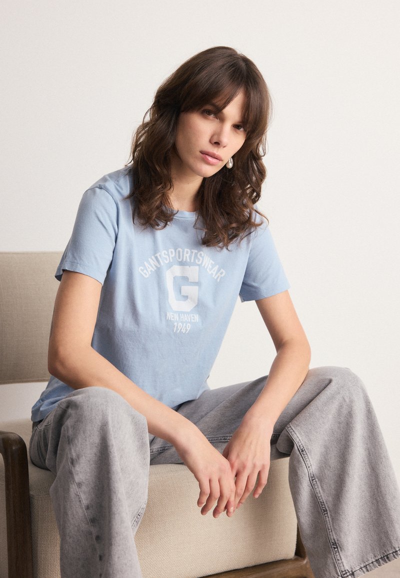 Kvinna med vågigt brunt hår som bär en ljusblå T-shirt med logotyp och grå vida jeans, sittande på en beige vadderad stol mot en vit vägg.