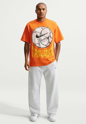 Mand stående iført en orange t-shirt med et stort motiv af en fodbold og pigtråd, grå joggingbukser og hvide sneakers.