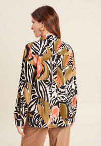 Chemise à manches longues avec un motif floral et de feuilles audacieux en noir, blanc, orange et vert. Conçue en tissu léger, elle présente un détail boutonné au niveau des poignets.