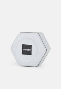 Conteneur en métal hexagonal de couleur gris clair avec un logo G-SHOCK noir. Présente un texte en relief, une surface lisse et des accents métalliques.