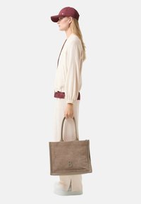 Beżowa torebka z przeszyciami, z wytłoczonym logo, miękka w dotyku, prostokątna, z podwójnymi uchwytami, zestawiona z jasnym strojem i czapką.
