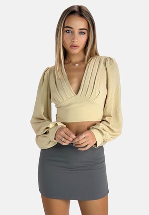 Blouse beige courte avec un décolleté en V profond et un design plissé, dotée de manches longues évasées. Associée à une mini-jupe grise ajustée.