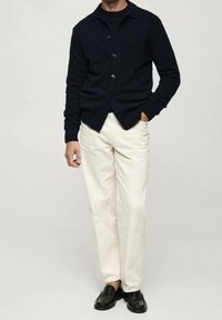 Dunkel marineblaue Strickjacke mit Knöpfen, getragen über einem schwarzen Hemd, kombiniert mit cremefarbenen Hosen und schwarzen Lederschuhen. Glatte Textur, tailliertes Design.